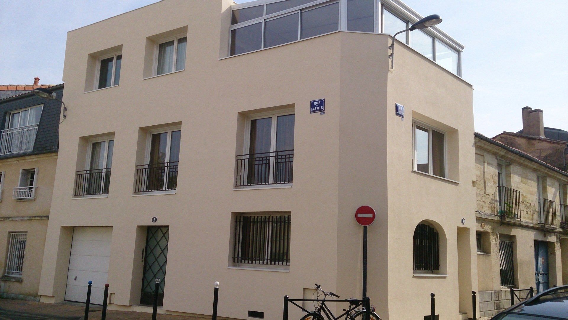 Un immeuble de trois étages, de couleur beige, situé à un angle de rue, avec des fenêtres rectangulaires.