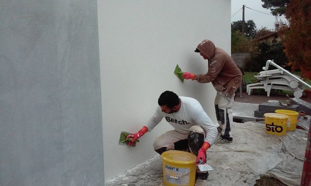 Deux ouvriers en tenue de protection appliquent une finition blanche sur un mur extérieur.