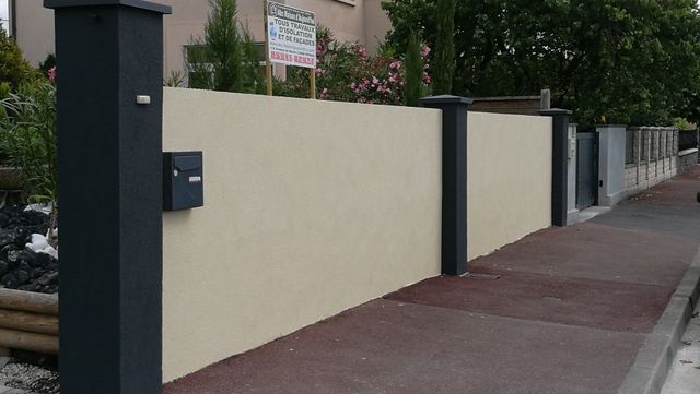 Un mur de clôture de couleur beige récemment rénové, avec des poteaux gris foncé et une boîte aux lettres assortie.