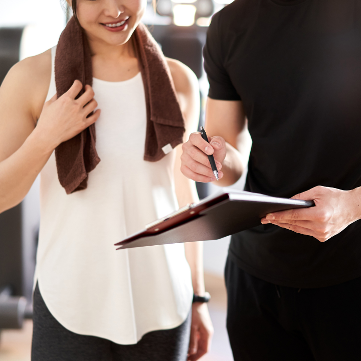 Mujer en el gimnasio con toalla, sonriendo, hablando con el entrenador que sostiene una carpeta y un bolígrafo.