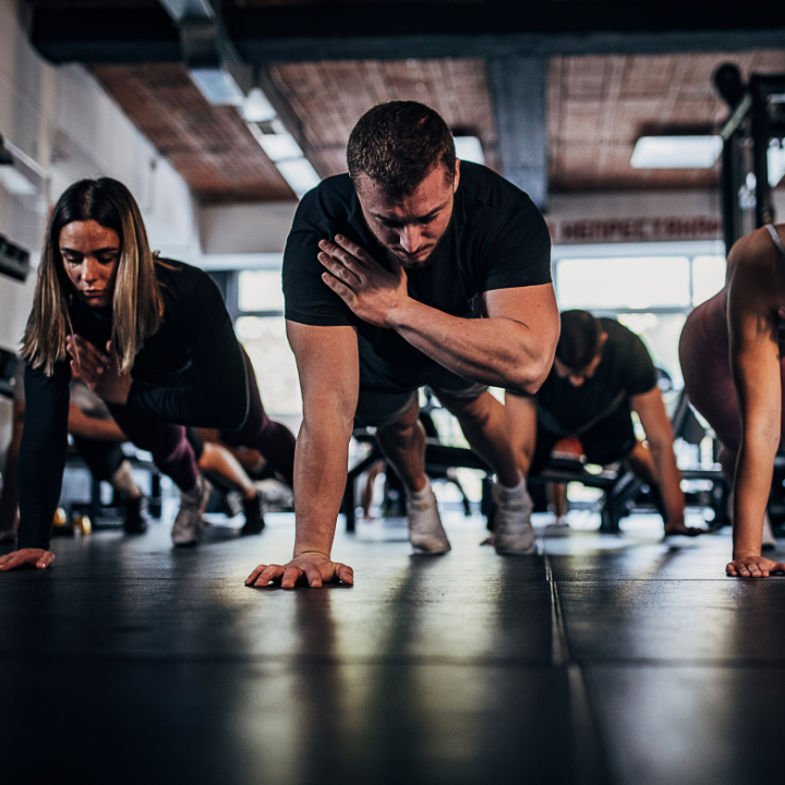 Personas haciendo flexiones en el gimnasio; un brazo levantado, concentradas en el ejercicio. Sala oscura con aparatos
