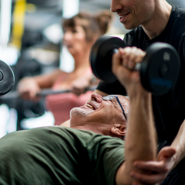 Un hombre realiza press de banca con mancuernas con la ayuda de un entrenador en un gimnasio. Al fondo, una mujer entrena 