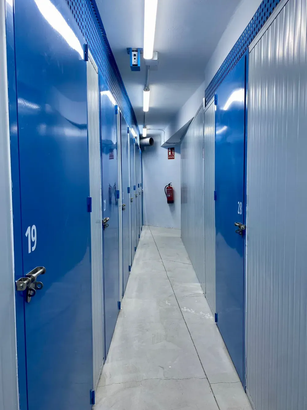 Un pasillo estrecho e iluminado en un almacén, con puertas azules numeradas a ambos lados y un extintor al final.