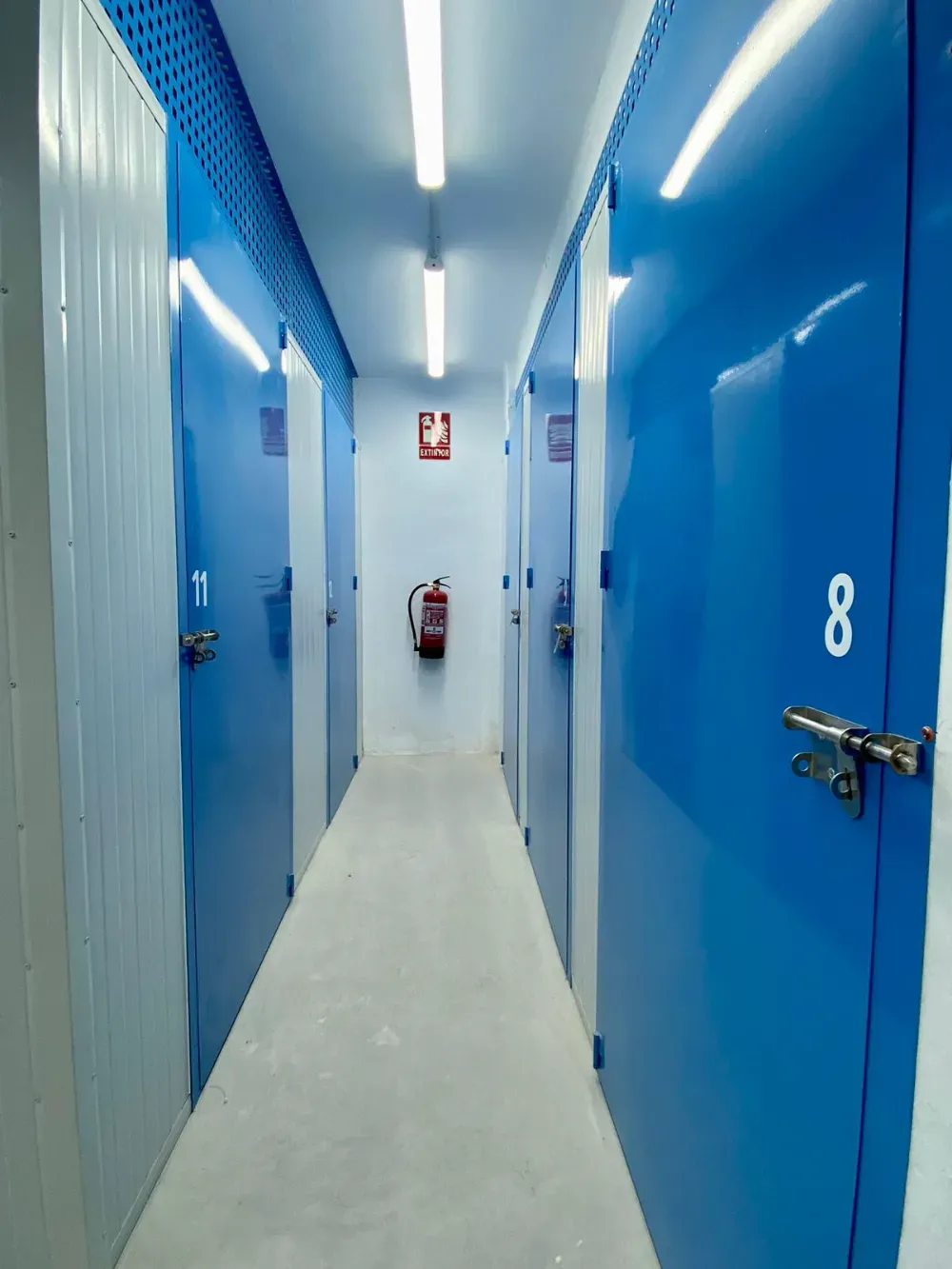 Un pasillo estrecho en un almacén de autoalmacenamiento con puertas numeradas de color azul y un extintor de incendios en la pared del fondo.