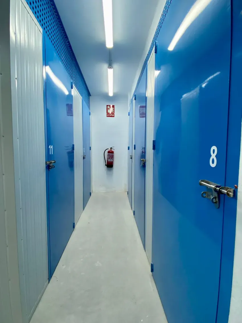 Un pasillo estrecho en un almacén de autoalmacenamiento con puertas numeradas de color azul y un extintor de incendios en la pared del fondo.