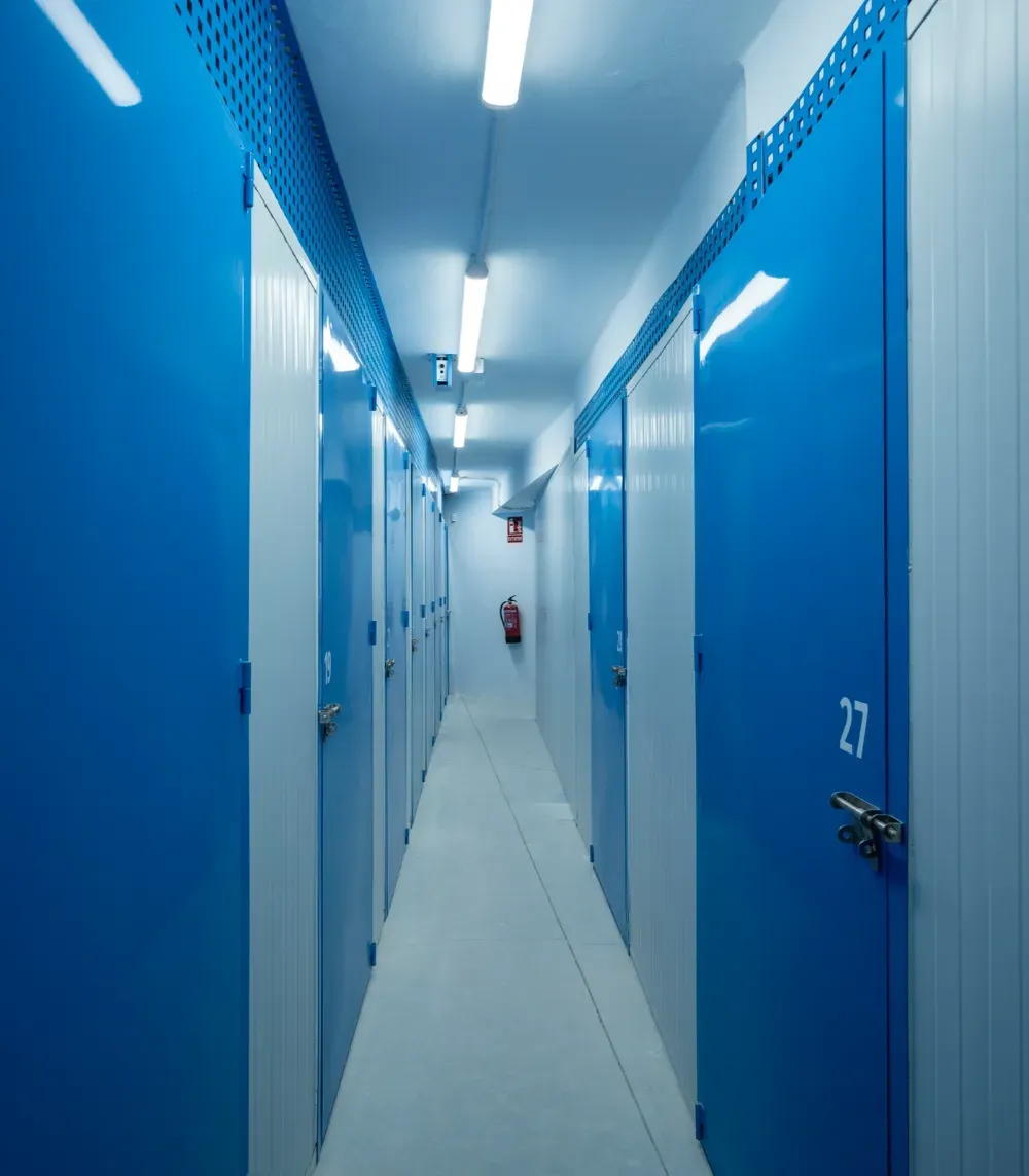 Un pasillo estrecho y bien iluminado, con puertas de trasteros azules y blancas, donde se ve claramente la puerta número 27.