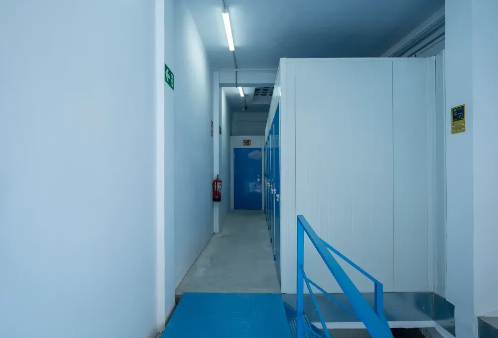 Un pasillo estrecho y luminoso con paredes blancas que conduce a una puerta azul, con una barandilla metálica azul en primer plano.