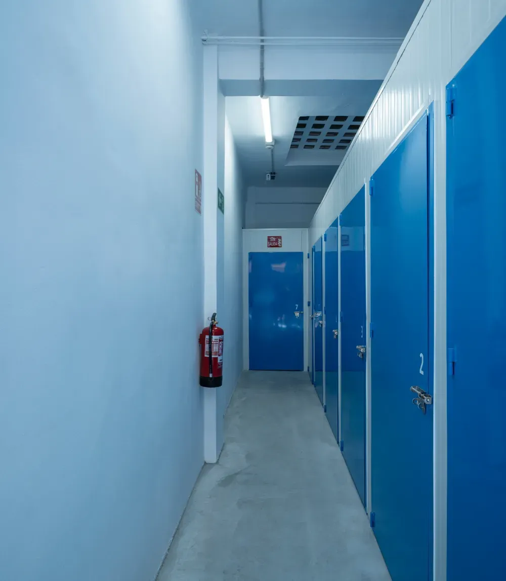 Un pasillo estrecho con paredes de color azul claro y suelo de hormigón, con una hilera de puertas de almacenamiento azules y un extintor de incendios.