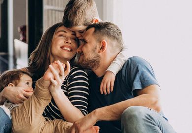Una familia de cuatro personas se abraza fuertemente en un sofá, sonriendo en un cálido momento en el interior.