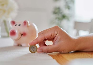 Una mano coloca una moneda junto a una hucha rosa sobre una mesa de madera.