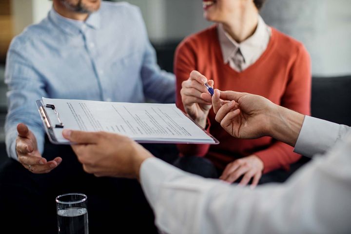 Reunión de negocios con portapapeles y bolígrafo mientras dos personas discuten documentos sobre una mesa.