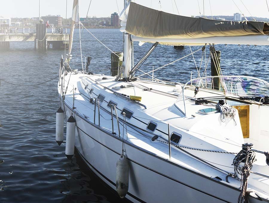 Un velero blanco atracado en aguas tranquilas, con los cabos del muelle y el puerto de la ciudad al fondo.