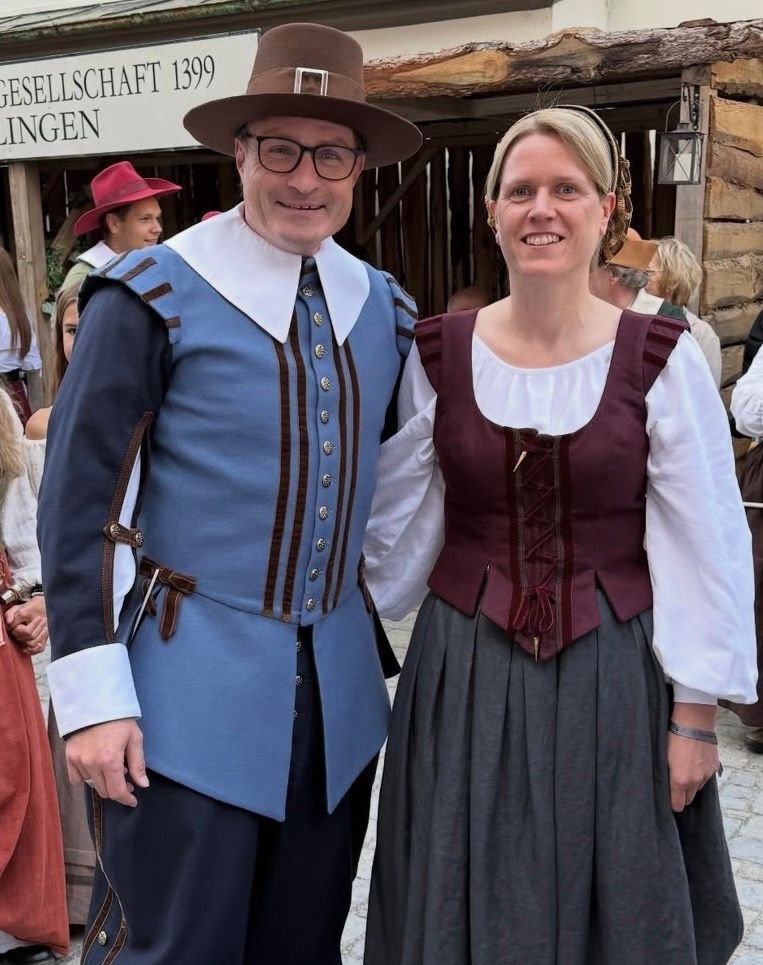Zwei Personen in historischer Kleidung posieren vor einem Gebäude; der Mann in Blau, die Frau in Burgunderrot.