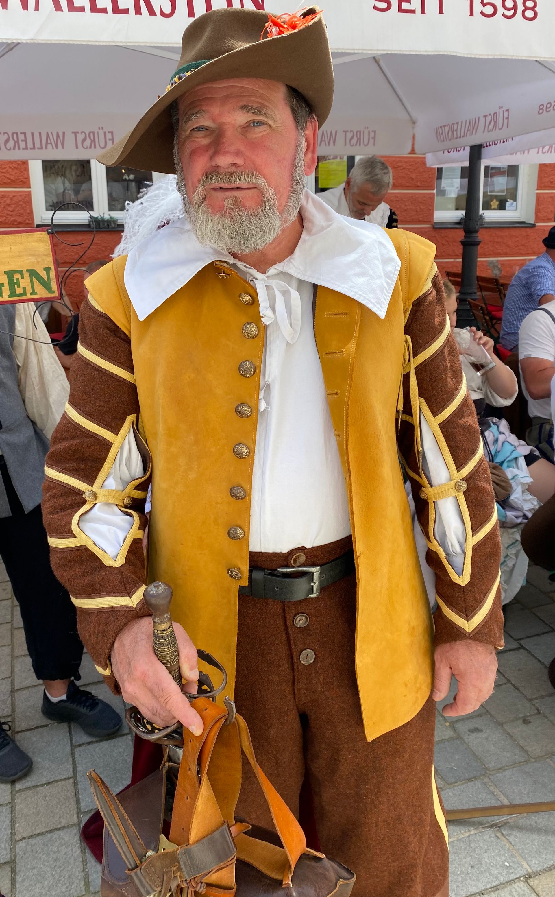 Mann in historischer Tracht, braun-goldenes Outfit, hält ein Schwert und lächelt im Freien.