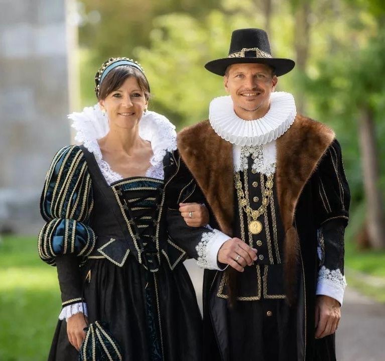 Ein Paar in historischer Kleidung mit Halskrausen, schwarzen Mänteln und Hüten posiert im Freien.