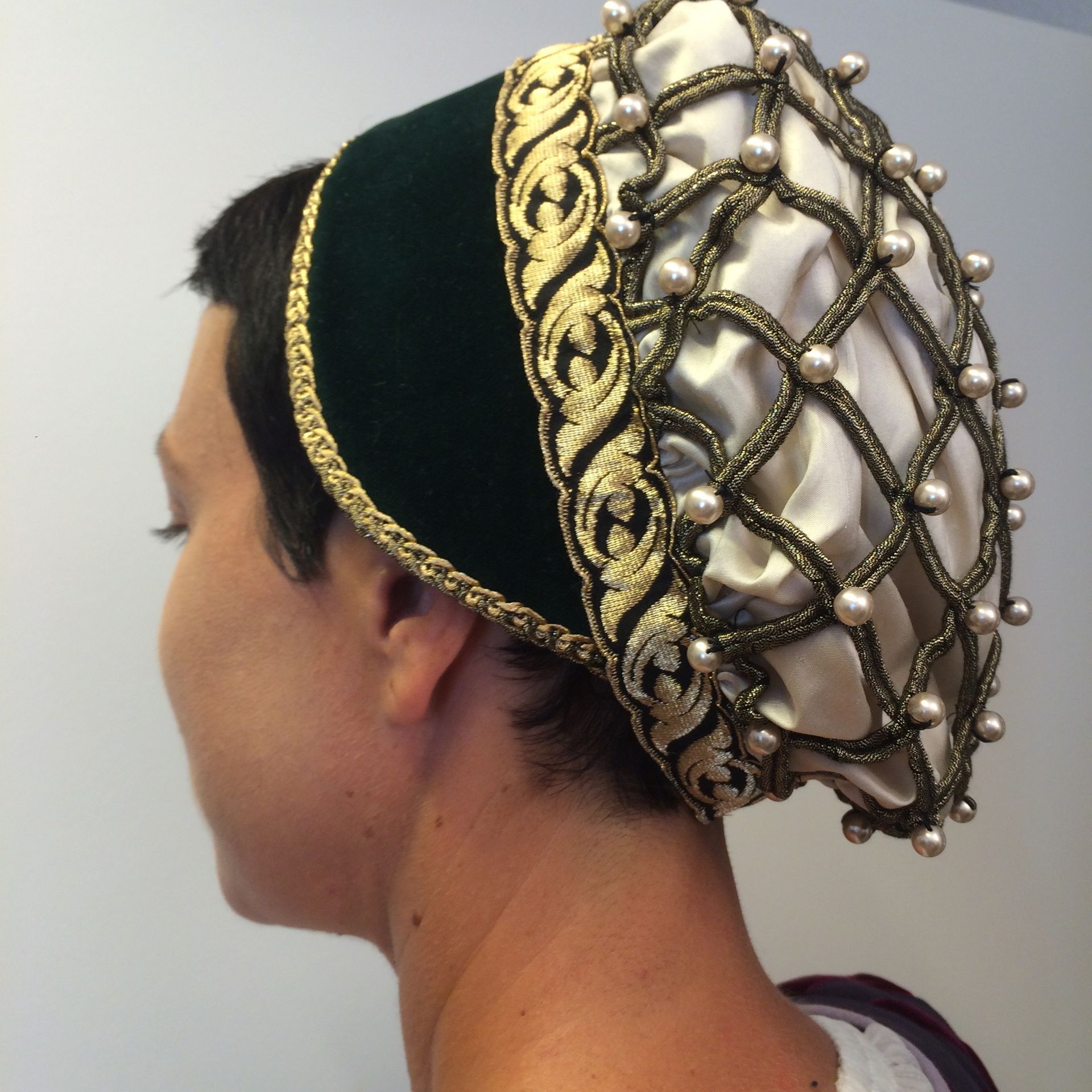 Person mit einem dunkelgrünen Samthut mit Goldrand, einer perlenbesetzten Netzhaube und weißem Satinfutter.