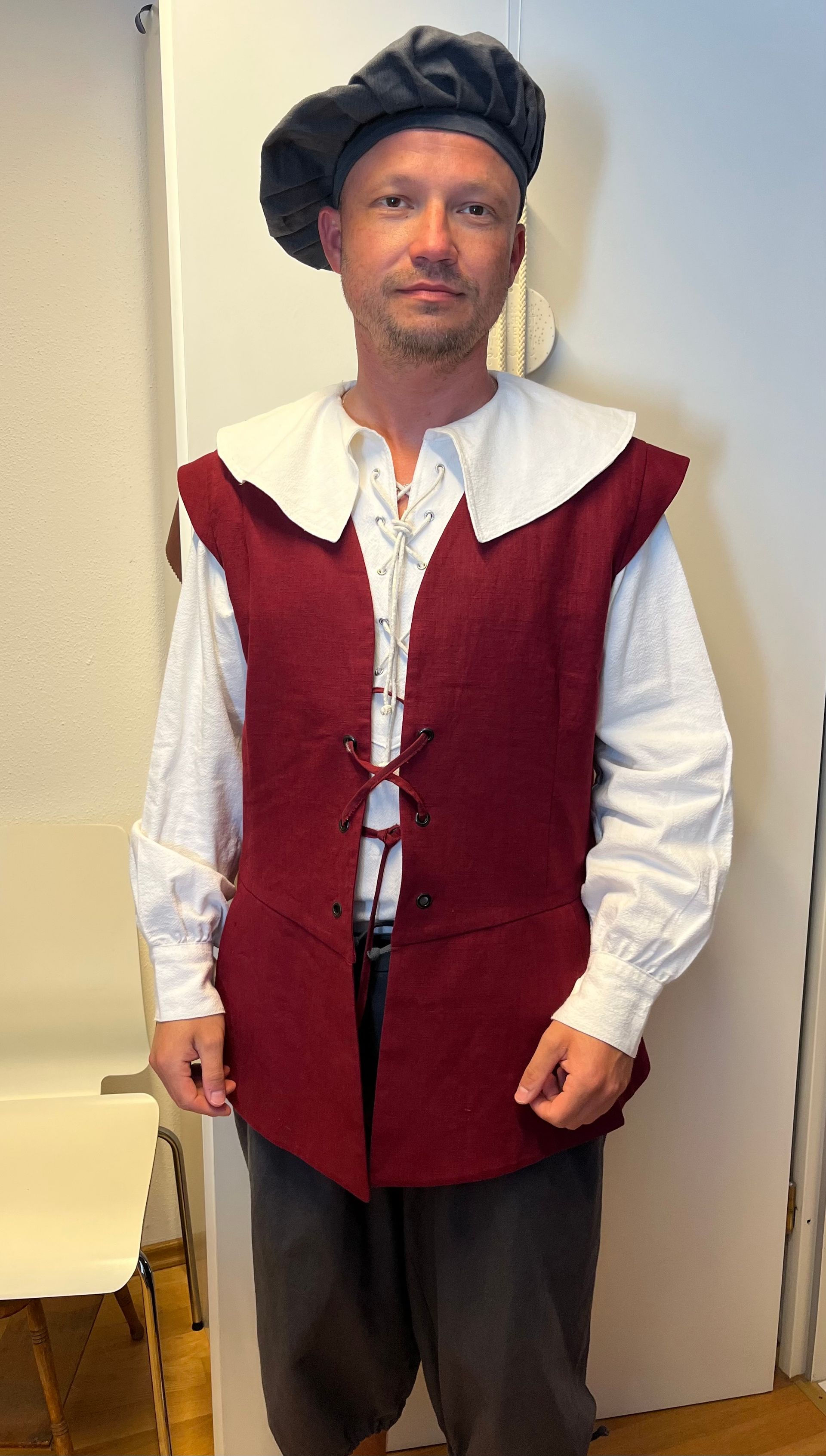 Ein Mann in bordeauxroter Weste und weißem Kragen, der einen Hut und ein helles Hemd trägt, steht.