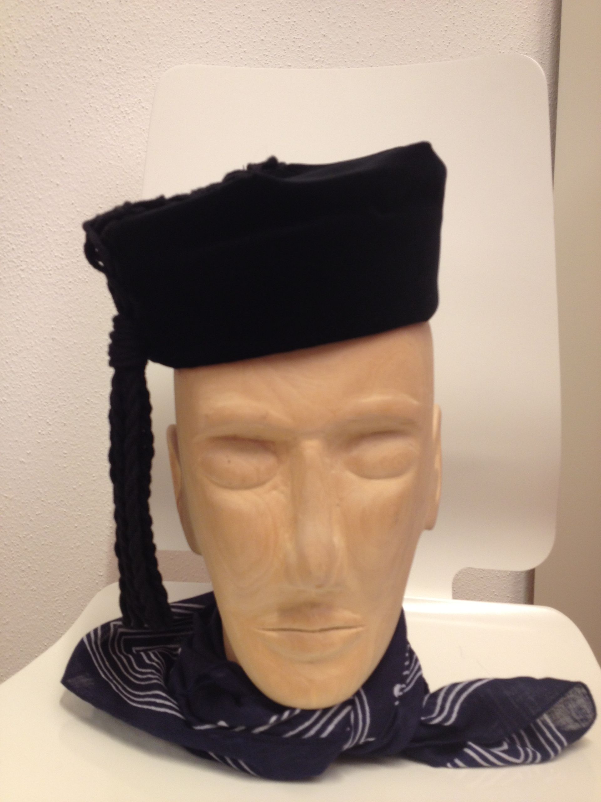 Auf einem hölzernen Schaufensterpuppenkopf sitzt ein schwarzer Hut mit Quaste, um den Hals trägt er einen gemusterten Schal.