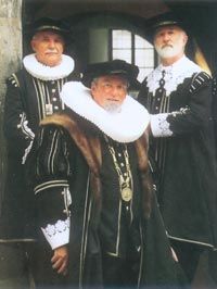 Drei Männer in dunklen Gewändern, weißen Halskrausen und Hüten posieren im Freien.