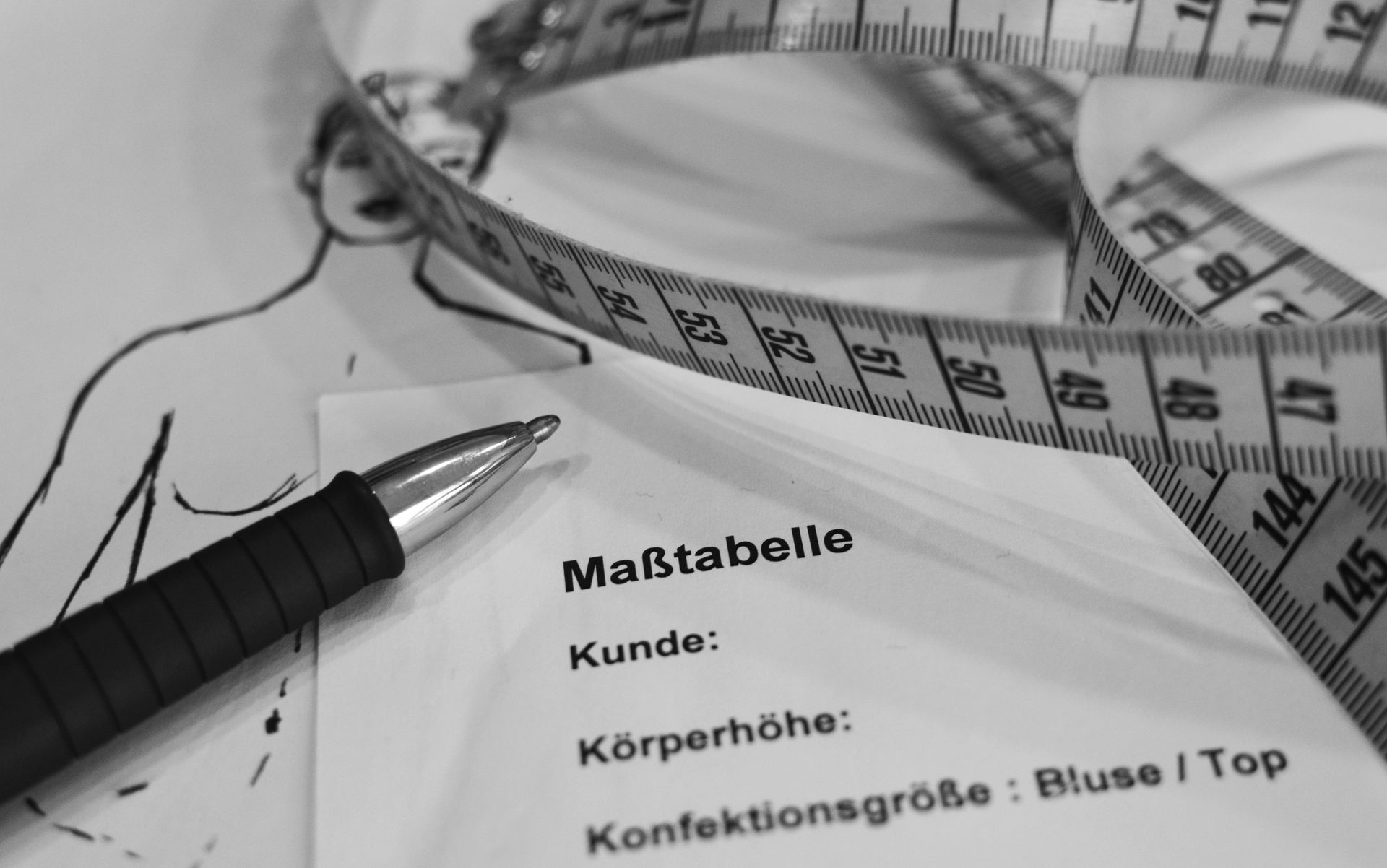 Eine Zeichnung einer Frau, eines Maßbandes, eines Stiftes und einer Maßtabelle für Kleidung in Schwarzweiß.