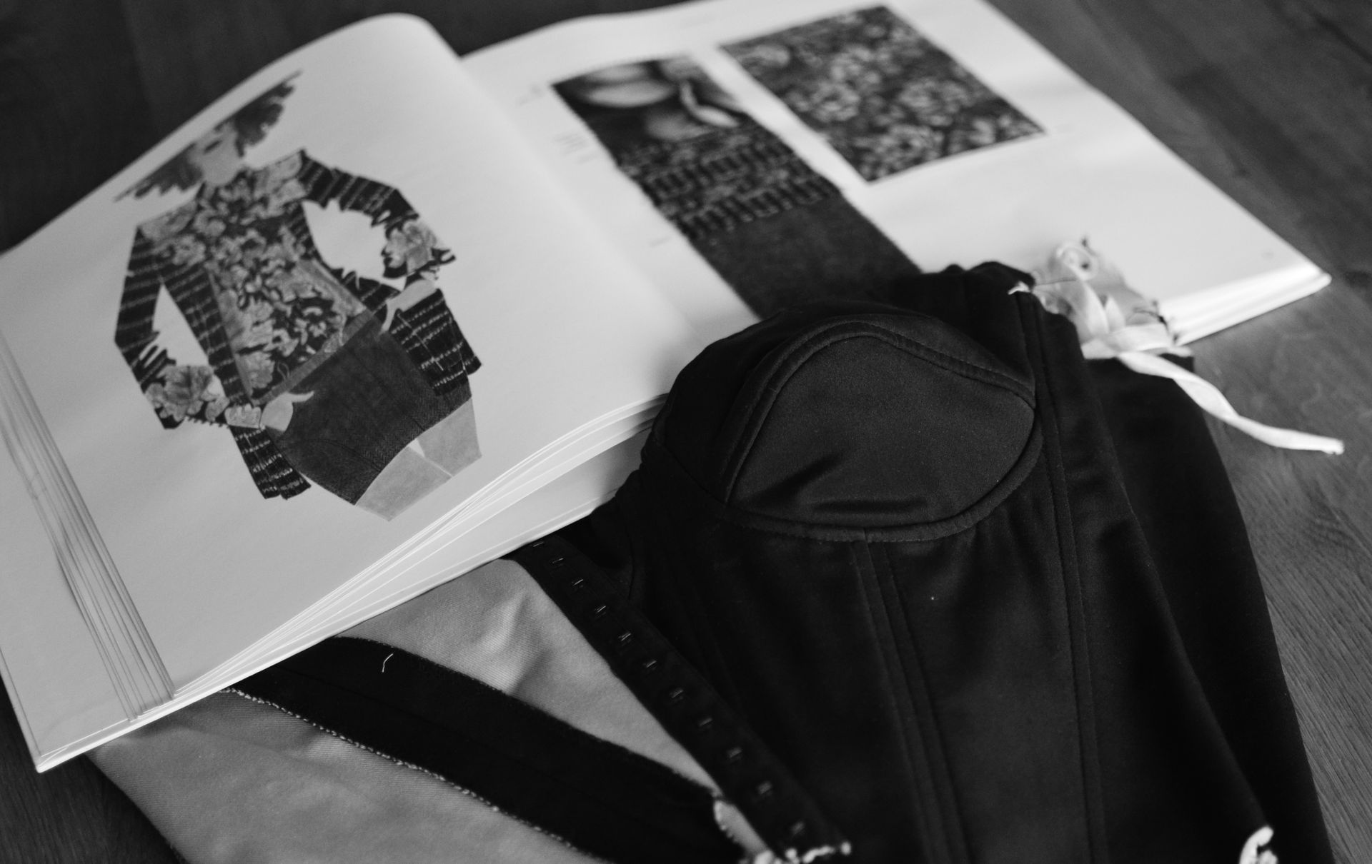 Schwarz-Weiß-Foto: Ein aufgeschlagenes Modedesignbuch neben einem schwarzen Korsett auf einer Holzoberfläche.
