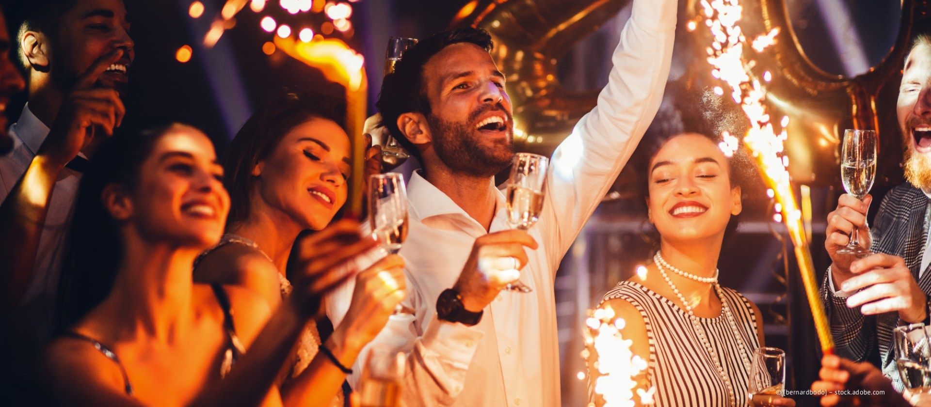 Menschen feiern, halten Champagnergläser und Wunderkerzen in den Händen und lächeln in einer schwach beleuchteten Umgebung.