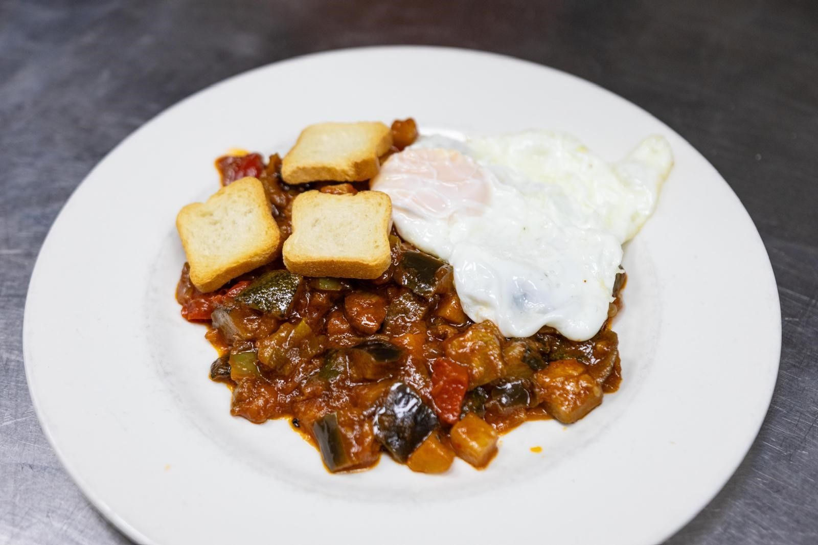 Una rebanada de pan tostado con tomate, un vaso de zumo de naranja y una taza de café humeante sobre una mesa de madera.