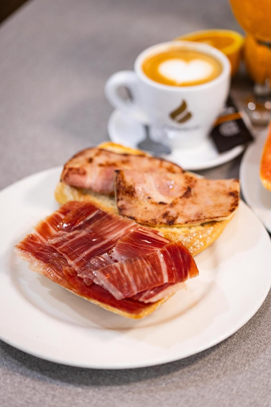 Un plato con tostada cubierta de jamón, junto a una taza de capuchino con arte latte y un vaso de zumo de naranja.