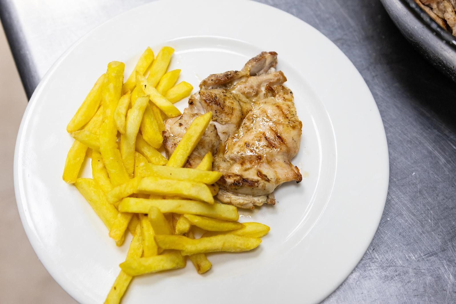 Un plato blanco con pechuga de pollo a la parrilla y una generosa porción de papas fritas doradas sobre una superficie gris.