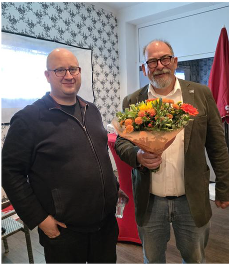 Zwei Männer, einer mit Glatze und Brille, der andere mit Bart, halten Blumen in der Hand und lächeln. Innenaufnahme, dekorative Tapete.