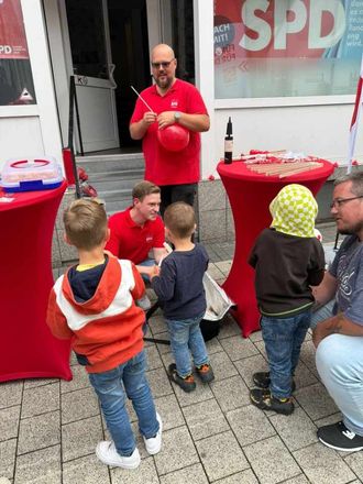 Menschen bei einer politischen Veranstaltung, die Kindern rote Luftballons geben. Vor einem Gebäude mit einem „SPD“-Schild.