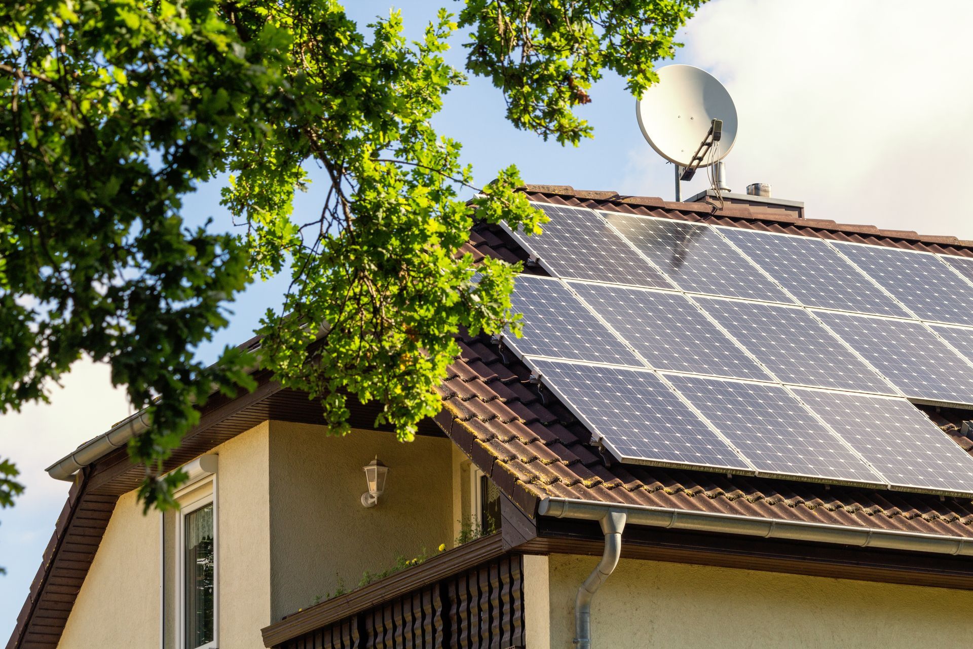 Toit de maison avec panneaux solaires et antenne parabolique à côté d'un arbre feuillu sous un ciel lumineux