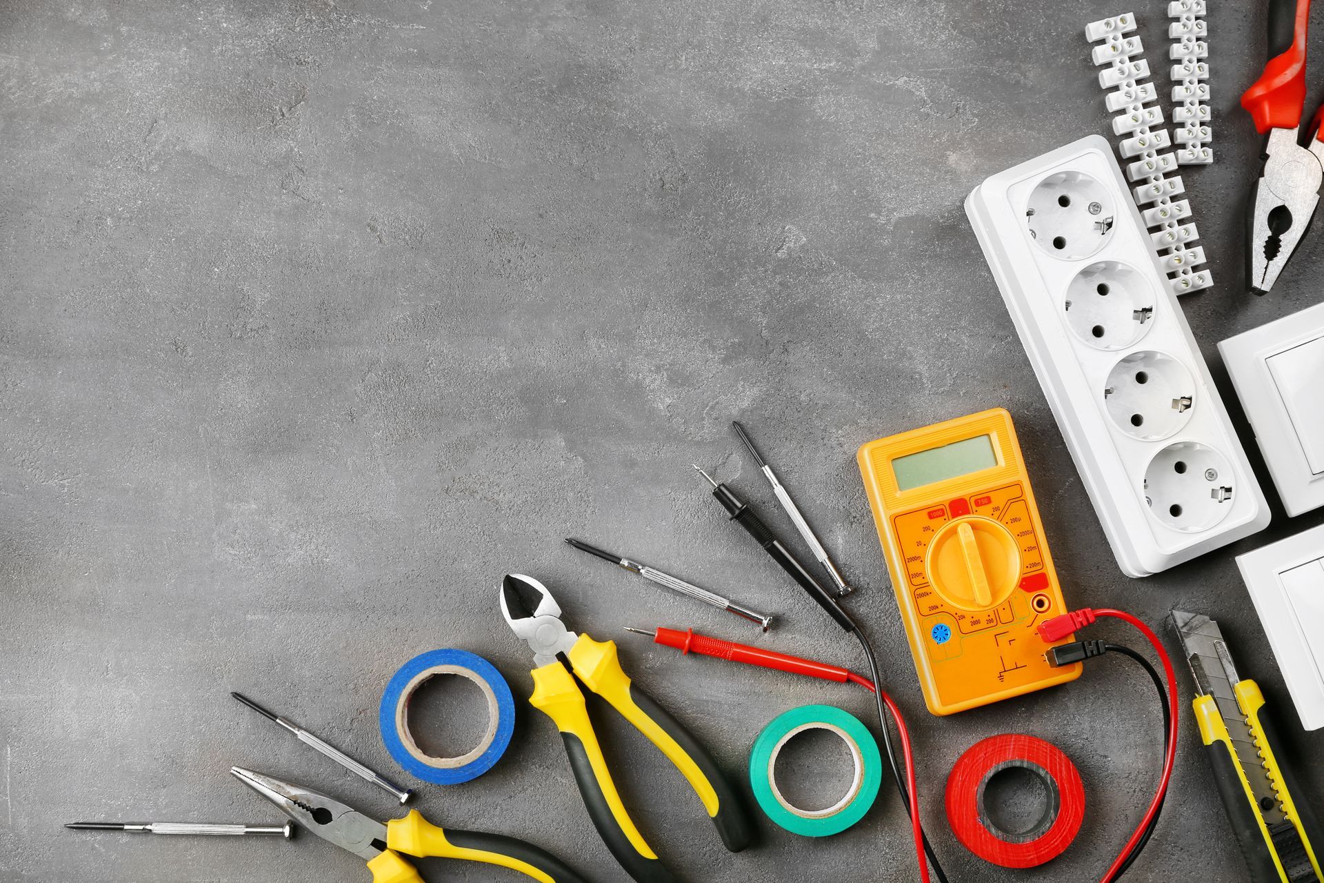 Outils électriques variés et multiprises sur une surface grise, comprenant des pinces, des tournevis, du ruban adhésif et des pinces coupantes.