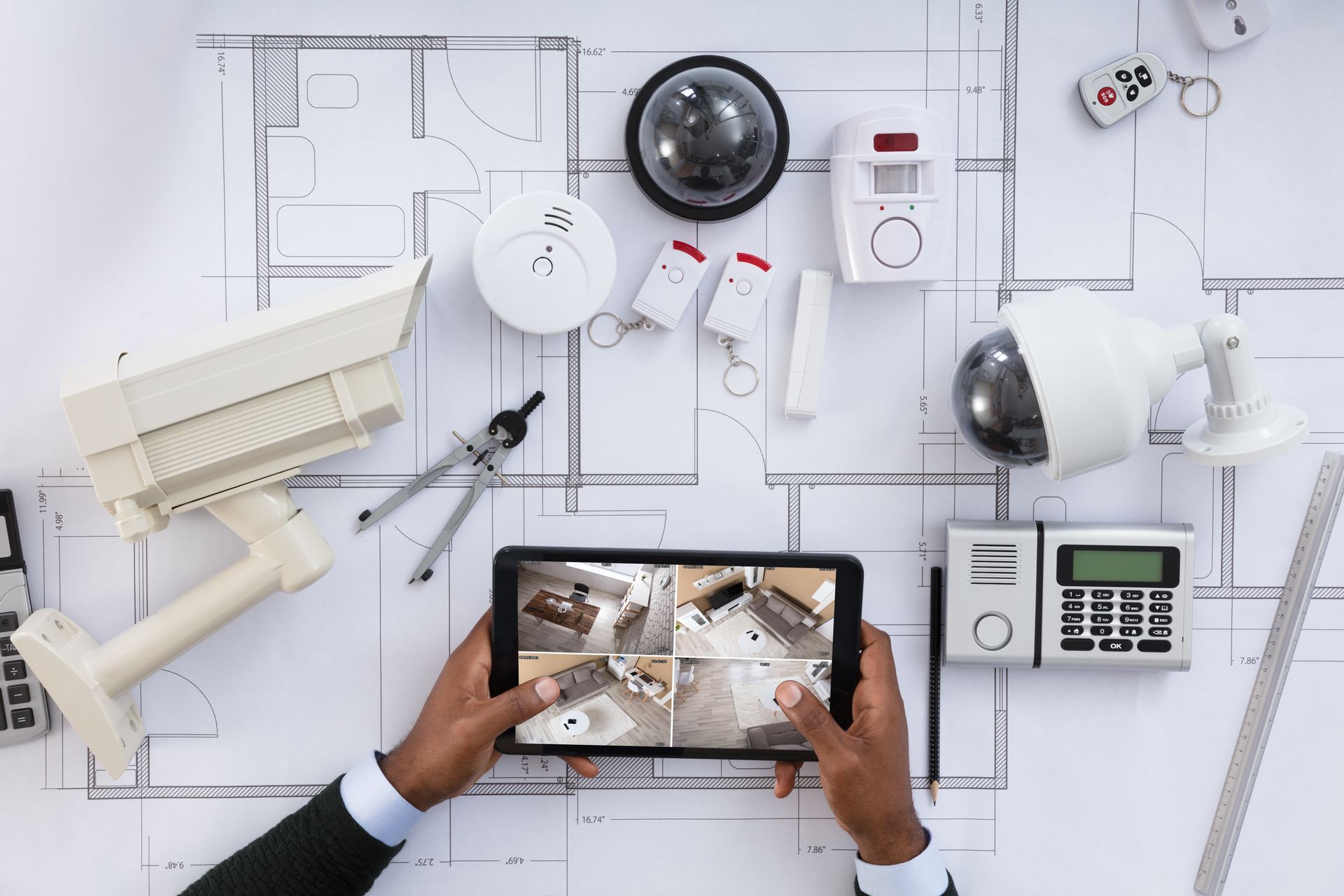 Des mains tenant une tablette affichant le plan d'une maison sont posées à côté d'un bureau blanc, à côté d'un bureau contenant du matériel et des outils de sécurité domestique.