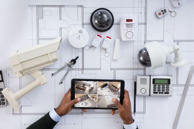 Des mains tenant une tablette affichant le plan d'une maison sont posées à côté d'un bureau blanc, à côté d'un bureau contenant du matériel et des outils de sécurité domestique.