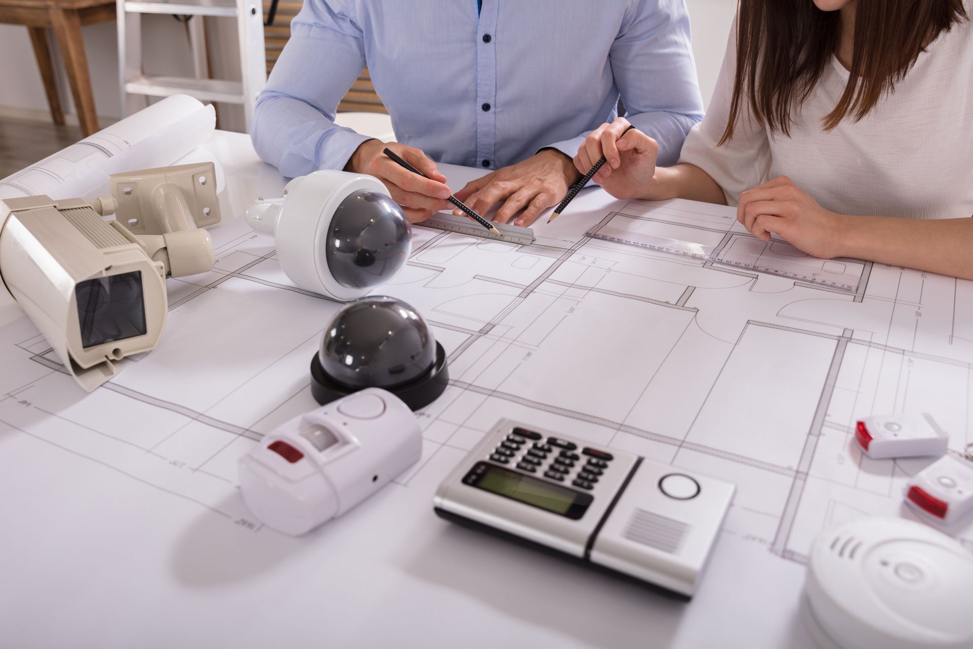 Deux personnes examinent les plans d'une maison avec des dispositifs de sécurité et une calculatrice sur la table.