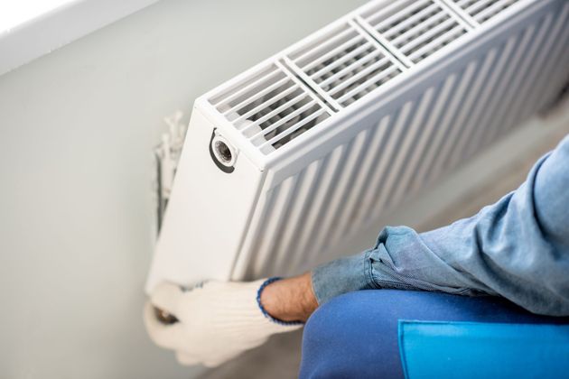 Une personne installant un radiateur blanc à l'aide d'un outil, portant une manche bleue et un jean