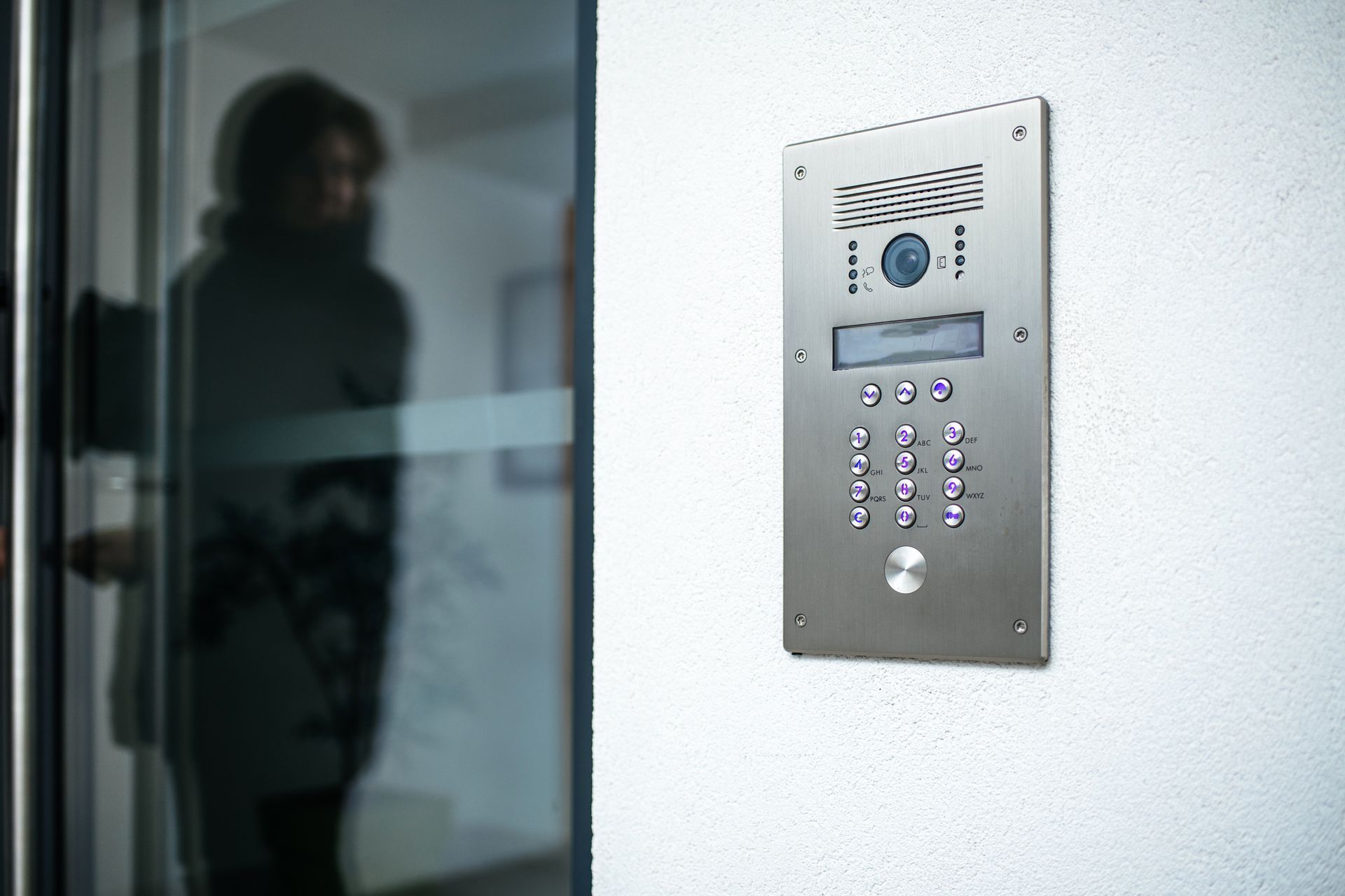 Panneau d'interphone à côté d'une porte vitrée, avec une silhouette floue reflétée dans le verre.