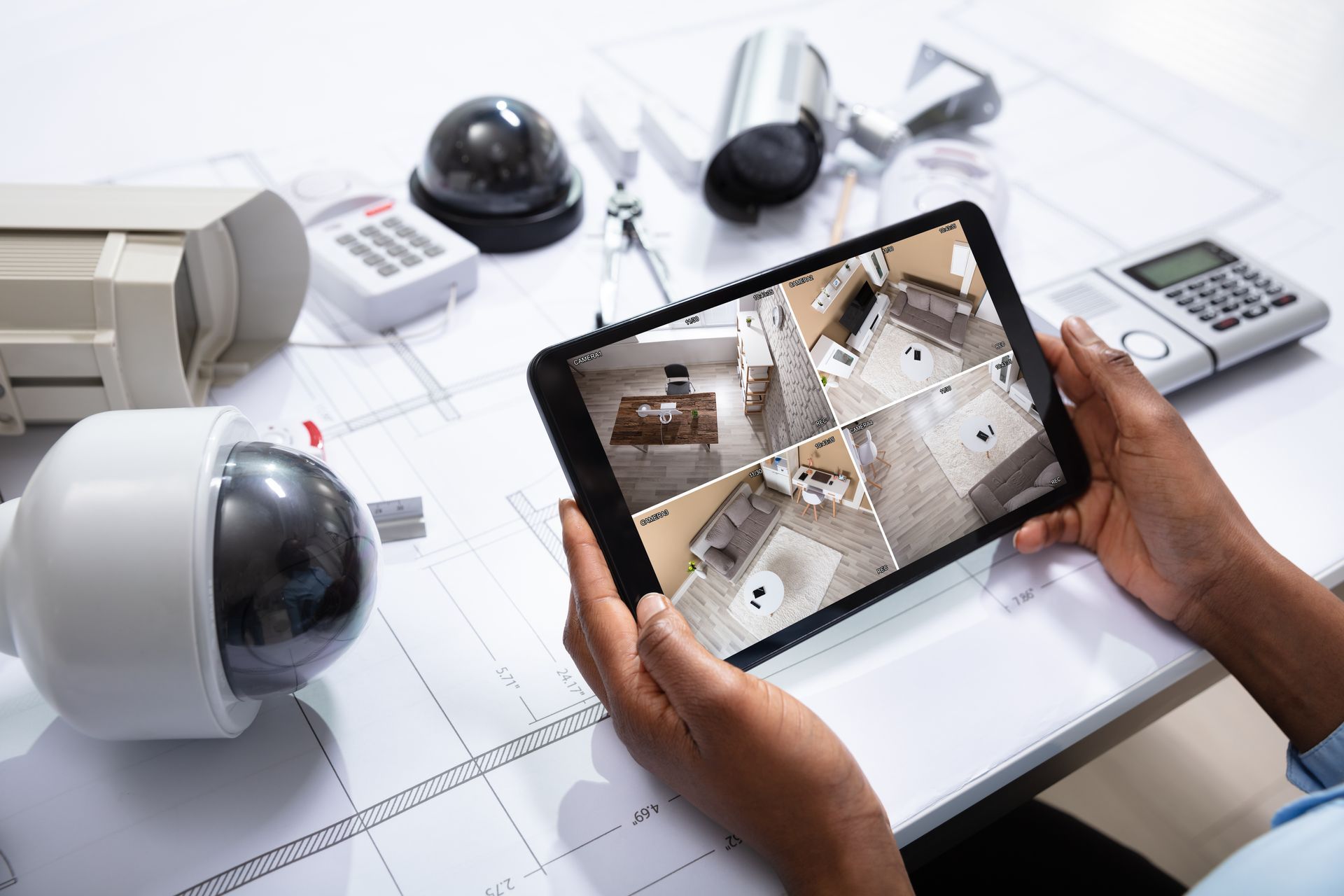Des mains tenant une tablette affichant les flux vidéo de caméras de sécurité, entourées de caméras de vidéosurveillance et d'équipements de surveillance sur un bureau