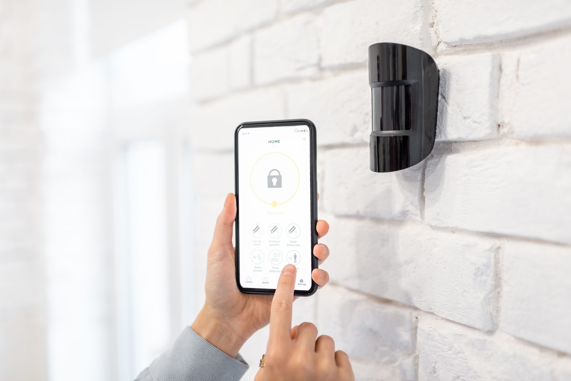 Une main utilise un smartphone pour contrôler une serrure connectée noire fixée au mur d'un mur de briques blanches.