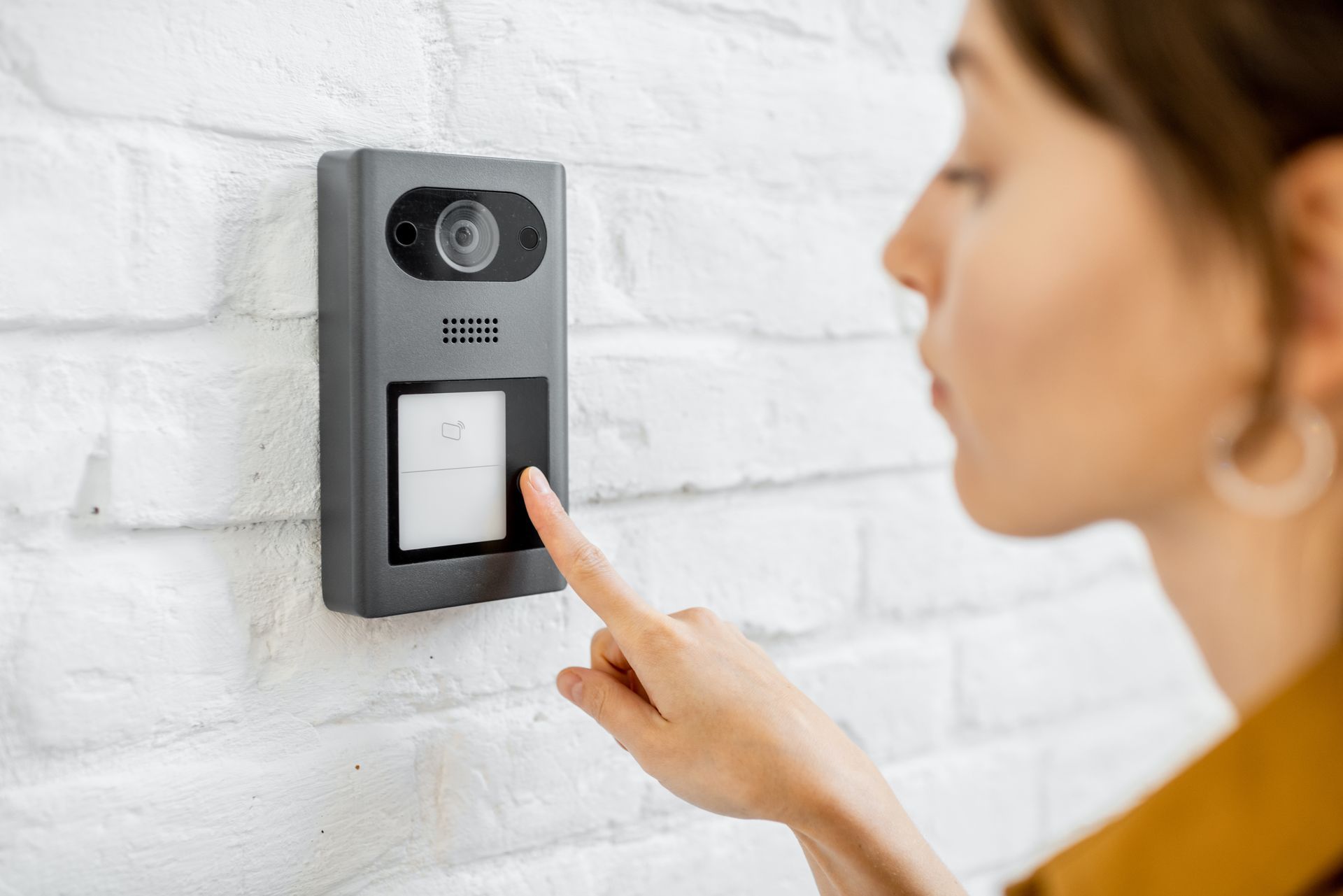 Une personne appuie sur une sonnette vidéo noire fixée sur un mur de briques blanches.