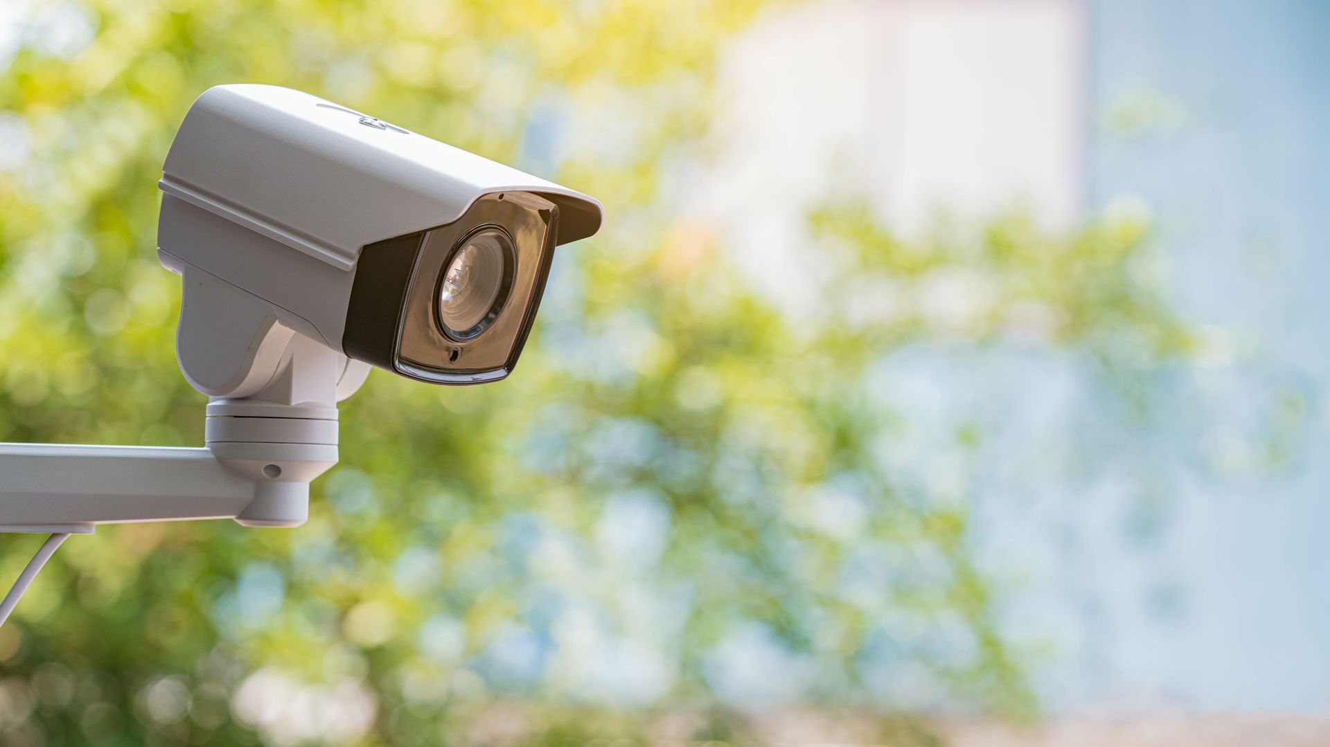 Caméra de sécurité extérieure blanche fixée au mur, pointée vers un jardin ensoleillé aux fleurs jaunes floues