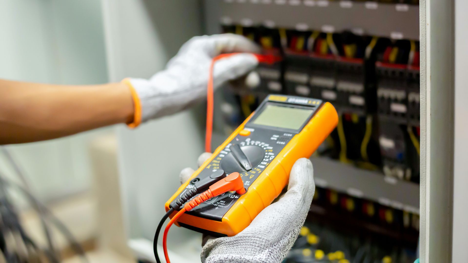 Des mains gantées utilisent un multimètre numérique pour tester le câblage électrique d'un panneau de commande.
