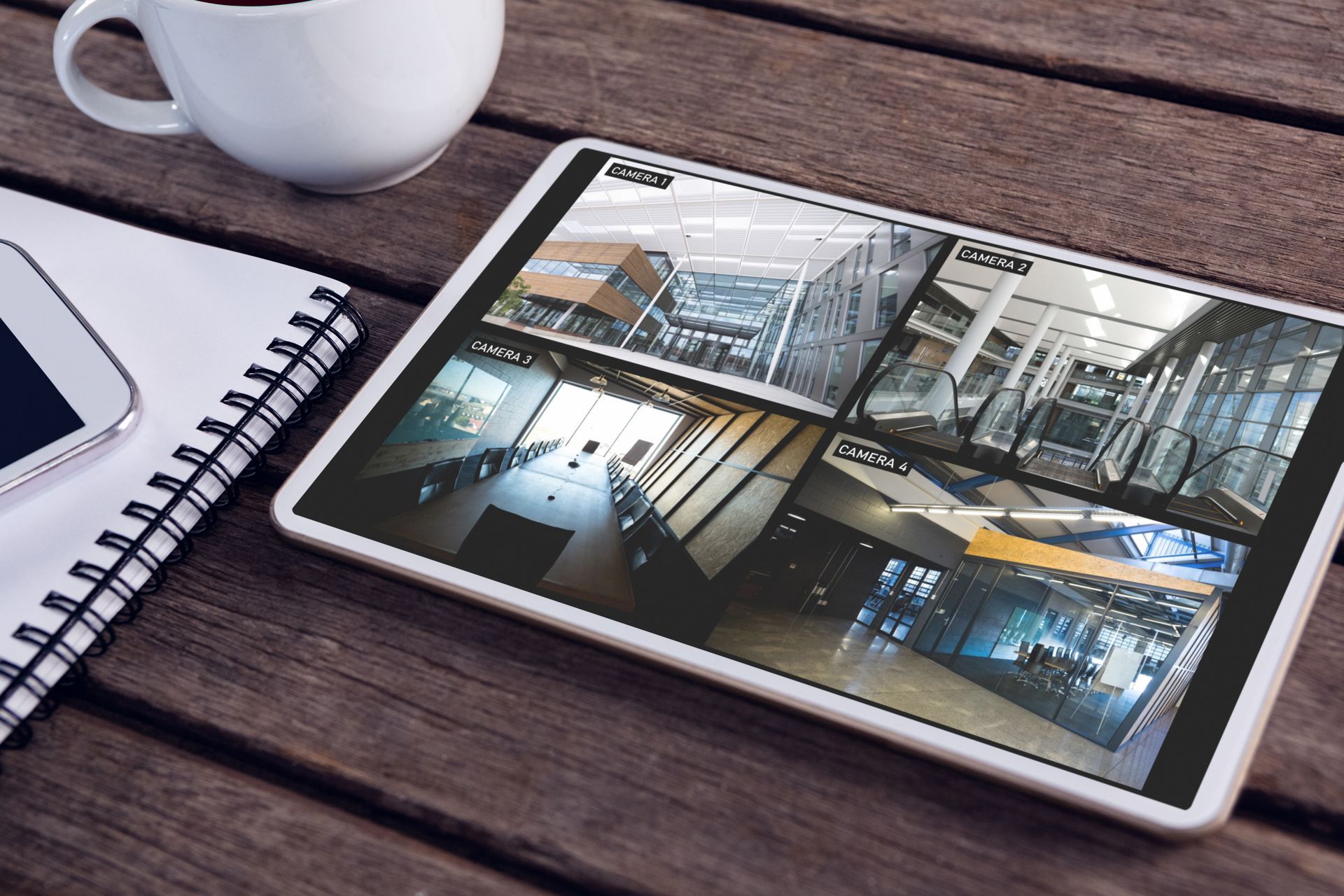Une tablette affichant les images d'une caméra de sécurité est posée sur un bureau en bois, à côté d'un ordinateur portable et d'une tasse à café.