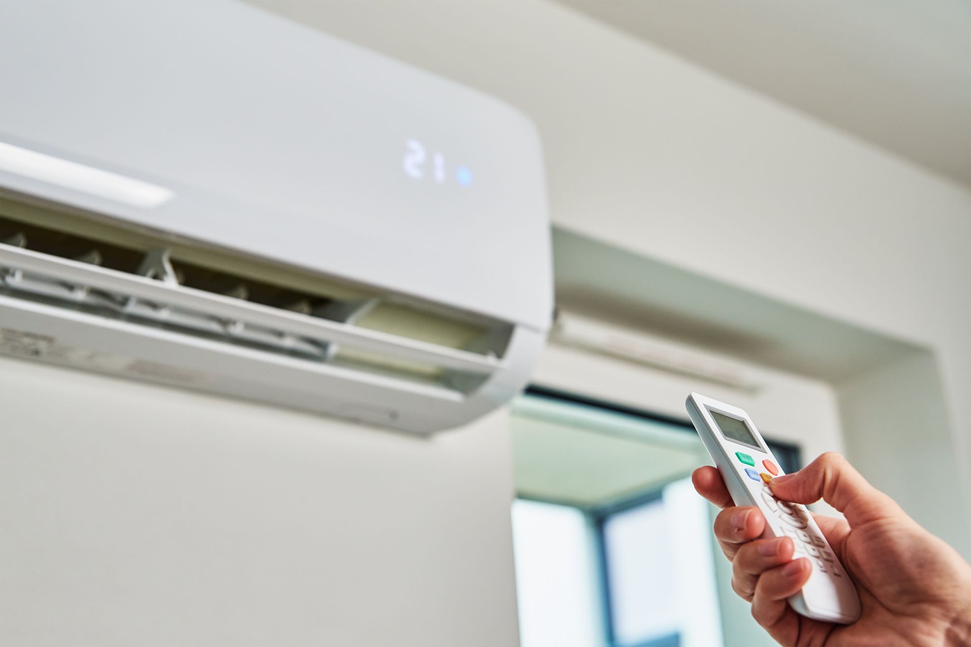 Main tenant une télécommande pointée vers un climatiseur mural dans une pièce lumineuse