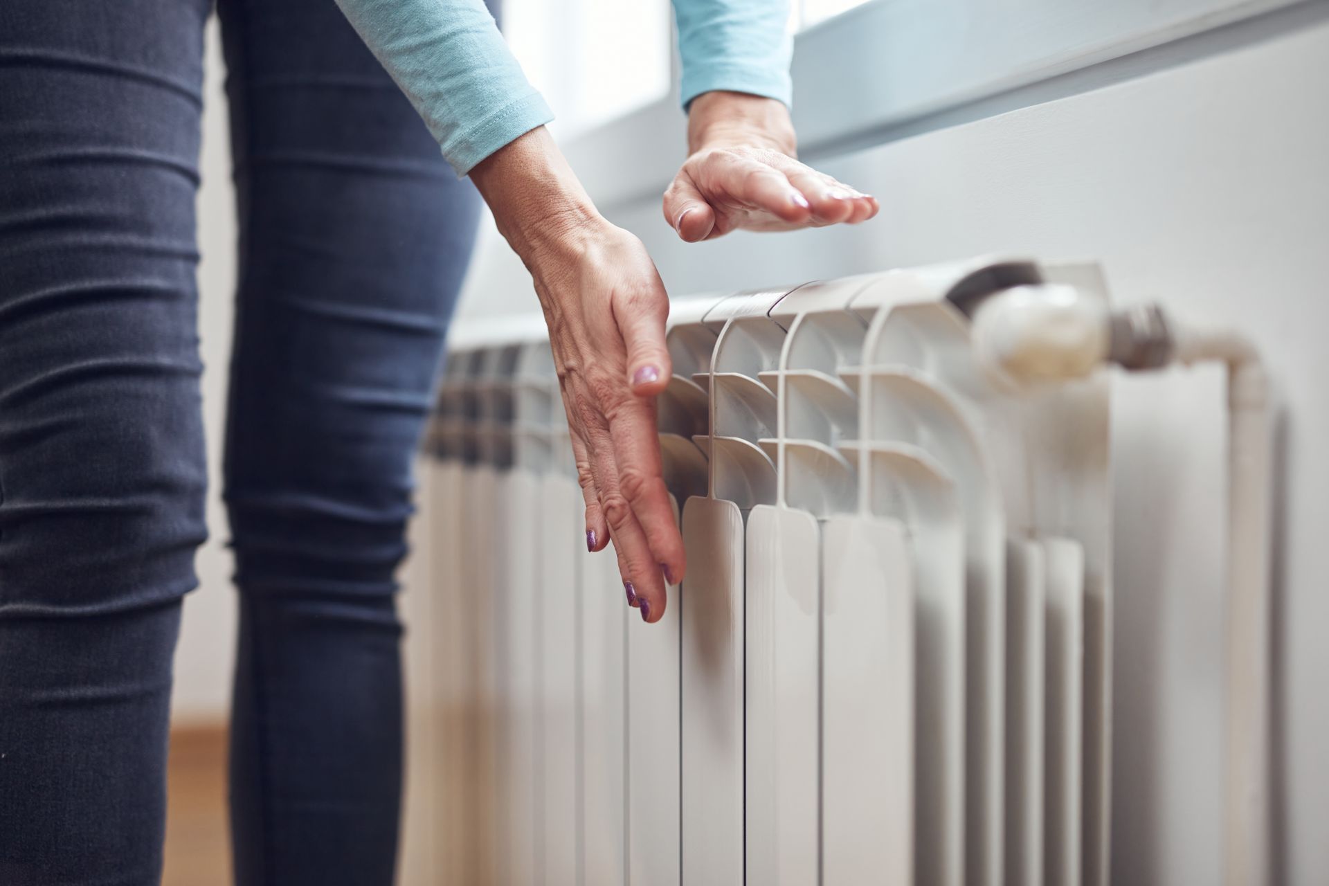Une personne en jean teste un radiateur blanc sous une fenêtre avec ses mains.