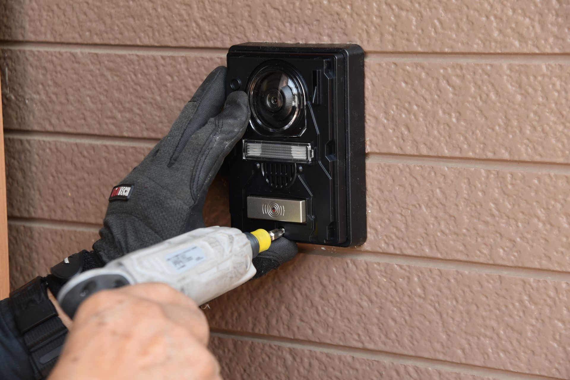 Des mains gantées utilisent une visseuse sur un cache-prise électrique extérieur noir fixé sur un bardage.