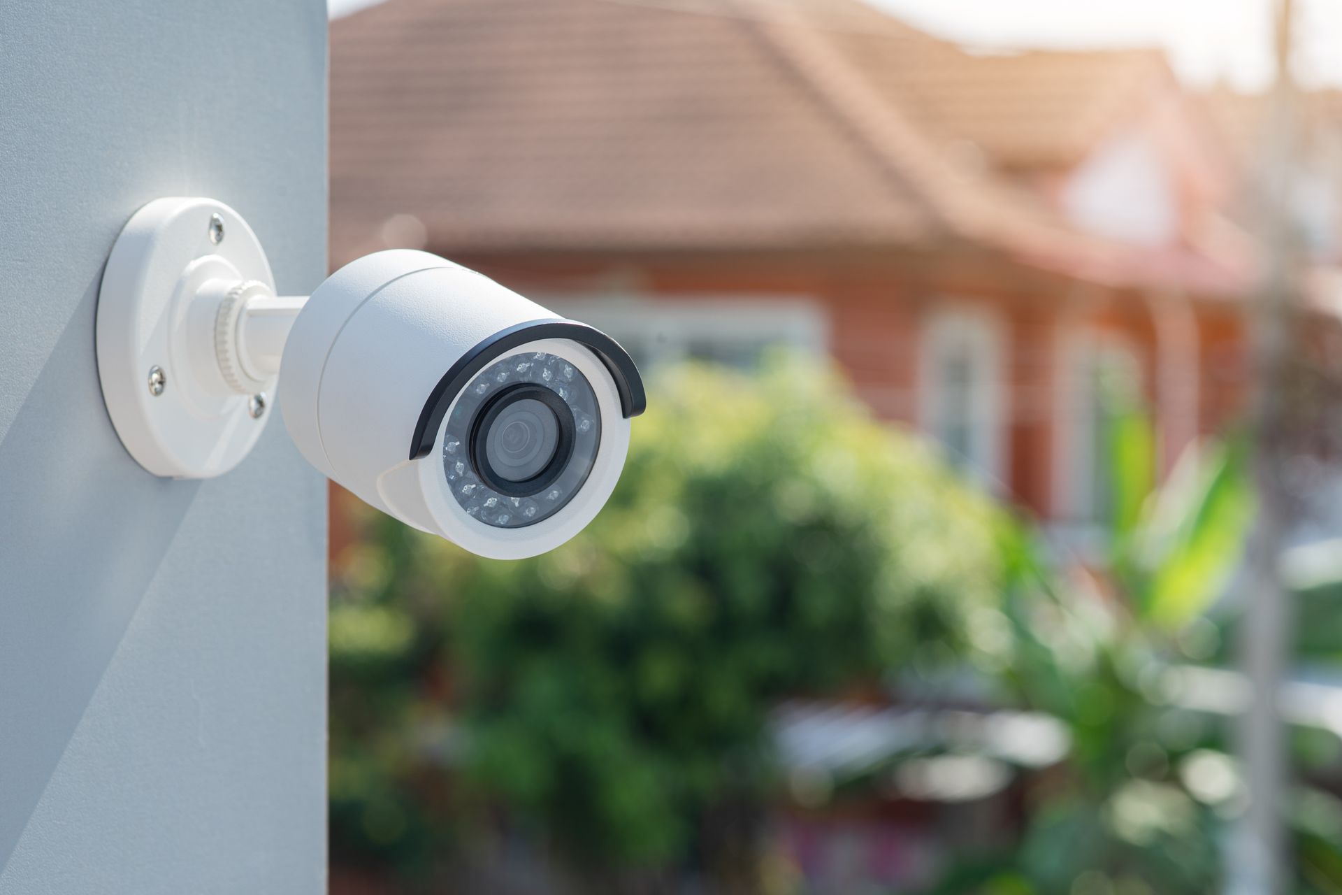 Caméra de sécurité blanche fixée sur un mur extérieur, donnant sur une maison et un jardin flous.