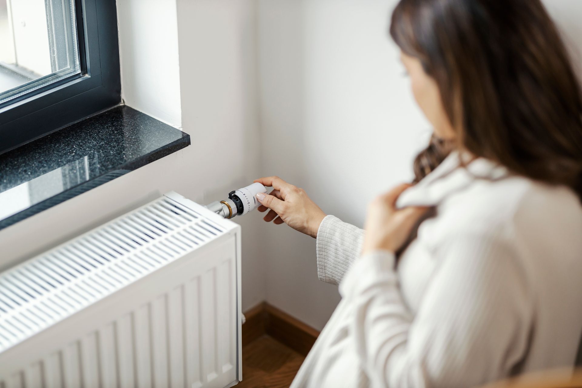 Une femme ajuste le thermostat d'un radiateur blanc près d'une fenêtre.
