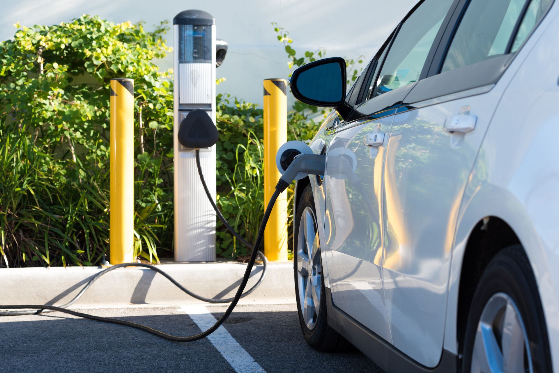 Voiture électrique blanche en charge à une borne publique extérieure, avec des bornes jaunes et de la verdure à proximité.